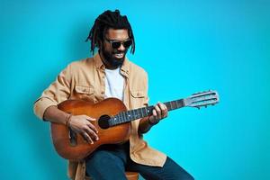 encantador joven africano con ropa informal tocando la guitarra y sonriendo mientras se sienta en un fondo azul foto