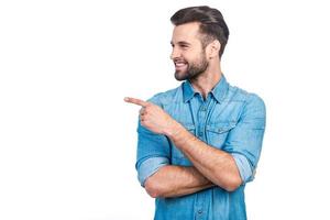 Look at that Happy young handsome man in jeans shirt pointing away and smiling while standing against white background photo