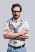 Confidence and style. Handsome young man keeping arms crossed and looking at camera while standing against grey background photo