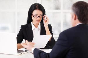 Job interview. Confident young woman in formalwear holding a paper and adjusting her glasses while grey hair man sitting in front of her and gesturing photo