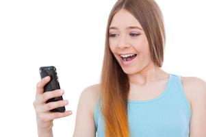 Message from him. Cheerful teenage girl holding mobile phone and looking at it with smile while standing isolated on white photo