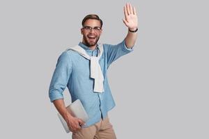 Good looking young man in eyeglasses holding his digital tablet and gesturing while standing against grey background photo