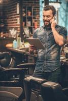 our appointment is on Monday  Cheerful young bearded man talking on mobile phone and looking at digital tablet while standing at barbershop photo