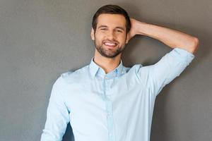 Feeling satisfied and relaxed. Handsome young man in shirt touching his head and smiling at camera while standing against grey background photo