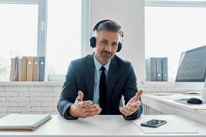 apuesto hombre de negocios con auriculares sonriendo y gesticulando mientras está sentado en su lugar de trabajo foto