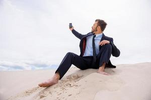 Poor signal. Frustrated young businessman searching for mobile phone signal while sitting on sand in desert photo