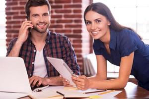 Keeping friendly atmosphere at work. Cheerful young man and woman sitting at the working place and smiling while woman holding digital tablet and man talking on the mobile phone photo