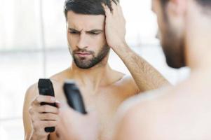 Bad shaver. Rear view of frustrated young man holding an electric shaver and looking at it while standing in front of the mirror photo
