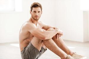 Rugged and handsome. Handsome young shirtless man in shorts sitting on the floor and looking at camera photo