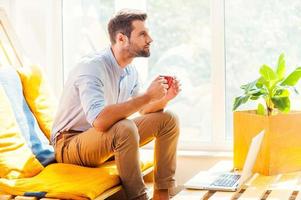 Coming up with new ideas. Side view of thoughtful young man holding cup of coffee and looking away while sitting in the rest area of the office photo