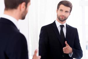 Young and successful. Handsome young man in full suit pointing himself and smiling while standing against mirror photo