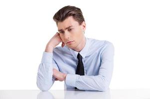 Feeling tired. Depressed young man in shirt and tie holding head in hand and looking away while sitting at the table and isolated on white photo