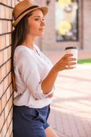 Relaxing with cup of fresh coffee. Side view of thoughtful young woman in funky hat holding cup with hot drink and looking away while leaning at the wall outdoors photo