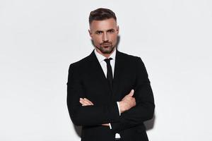 Confident business expert. Handsome young man in full suit keeping arms crossed and looking at camera while standing against white background photo