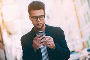 Quick message to friend. Handsome young man in smart casual wear holding smart phone and looking at it while walking along the street photo