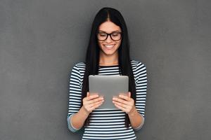 That is just great Attractive young woman holding digital tablet and looking at it with smile standing against grey background photo