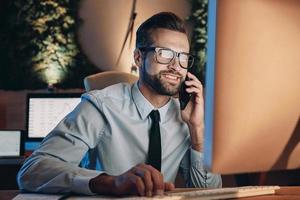 Cheerful young man working on computer and talking on mobile phone while staying late in the office photo
