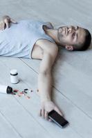 Too much pills. Top view of young man lying on the floor and holding a mobile phone in his hand while bottles with pills laying near him photo