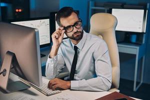 Confident young man adjusting his eyeglasses while staying late in the office photo