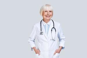 Healthcare worker. Beautiful senior woman in lab coat looking at camera and smiling while standing against grey background photo