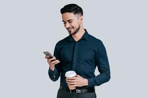 Nice chat. Good looking young man holding smart phone and looking at it with smile while standing against grey background photo