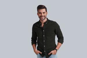 Young and successful. Good looking young man keeping hands in pockets and looking at camera with smile while standing against grey background photo