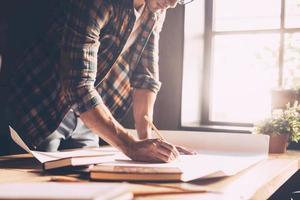 Working with passion. Close-up of confident young man in casual wear sketching on blueprint while standing near wooden desk in creative office photo