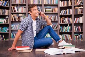 Talking with friend. Happy young man holding hands on chin and smiling while sitting against bookshelf photo