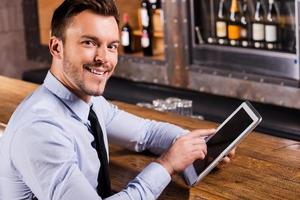 Businessman in bar. Handsome young man in shirt and tie working on digital tablet and smiling while sitting at the bar counter photo