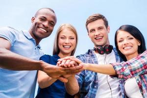 Together we are stronger. Low angle view of four happy young people bonding and holding hands clasped with blue sky in the background photo