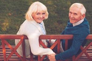 Happy to be together. Rear view of happy senior couple holding hands and looking at camera with smile while sitting on the park bench together photo