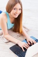 Surfing web at home. Top view of cheerful young teenage girl using computer and smiling while lying down on the floor at her apartment photo