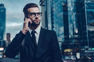 Business talk. Night time image of confident young man in full suit talking on the mobile phone and looking away while standing outdoors with cityscape in the background photo