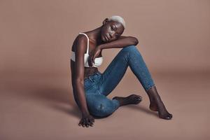 Confident in her beauty. Attractive young African woman looking at camera while sitting against brown background photo