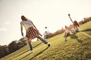 Supporting friends. Two young women sitting on the grass and gesturing while their male friends playing soccer outdoors photo