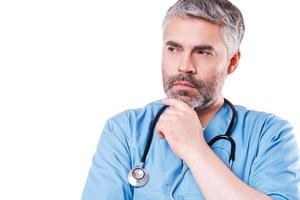 Thoughtful surgeon. Thoughtful mature grey hair doctor looking away and holding hand on chin while standing isolated on white photo