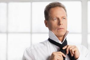 Morning dress up. Confident mature man in white shirt tying  his necktie and looking away photo