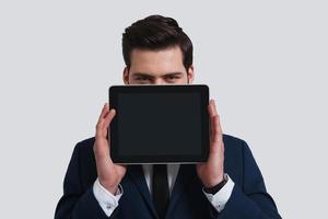 Using modern technologies. Good looking young man in full suit holding digital tablet and looking at camera while standing against grey background photo