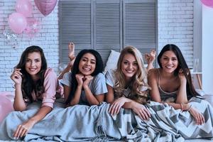 cuatro atractivas mujeres jóvenes en pijama sonriendo y mirando a la cámara mientras están acostadas en la cama foto