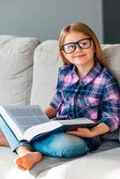 Little cutie with book. Cheerful little girl in glasses holding book and looking at camera with smile while sitting on the couch in lotus position at home photo