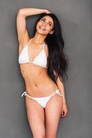 Fit and beautiful. Beautiful young mixed race woman in white bikini holding hand in hair and smiling while standing against grey background photo