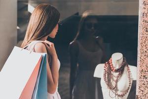 qué hermoso collar vista trasera de una hermosa joven que lleva bolsas de compras y mira a través de la ventana de la tienda de moda foto