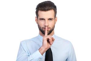 Keep silence  Confident young handsome man in shirt and tie holding finger on lips and looking at camera while standing against white background photo