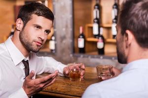 pasar la noche en el bar. dos jóvenes confiados con camisa y corbata hablando entre ellos y gesticulando mientras beben whisky en el mostrador del bar foto