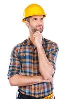 Thinking about new project. Thoughtful young male carpenter in hardhat holding hand on chin and looking away while standing against white background photo