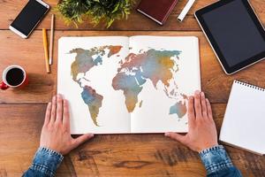 Planning his journey. Top view close-up image of man holding hands on his notebook with colorful map on it while sitting at the wooden desk photo