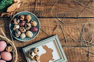 Easter table. Top view of Easter eggs on plates and Easter rabbit in picture frame lying on wooden rustic table with hay photo