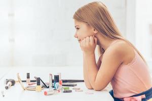 The most beautiful girl in the world. Side view of cheerful little girl keeping hands on chin and looking at her reflection in mirror while sitting at the dressing table photo