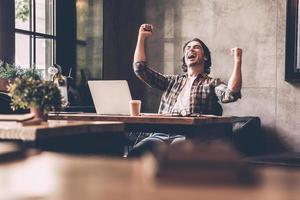 Everyday winner. Cheerful young man in casual wear keeping arms raised and looking happy while sitting at the desk in office photo