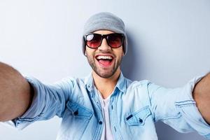 solo yo y nadie más. vista superior de un apuesto joven con sombrero y gafas de sol haciendo selfie y sonriendo mientras se enfrenta a un fondo gris foto